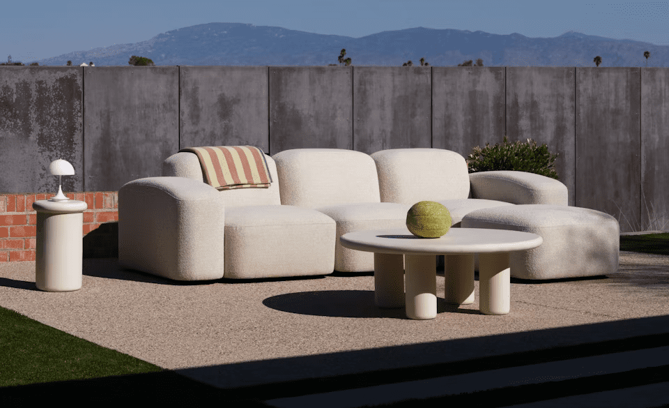 Cozy outdoor seating area featuring modern neutral-toned furniture, a round table with a decorative melon, and soft green grass, framed by a sleek gray wall and distant mountains.