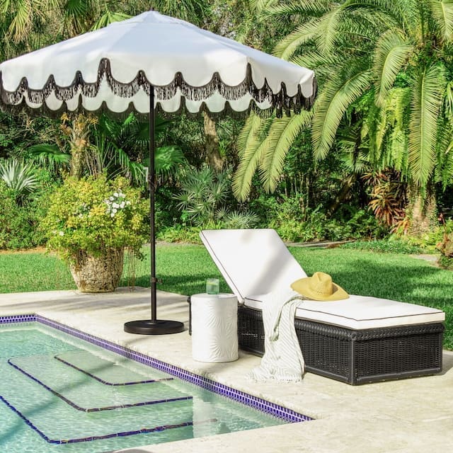 Luxurious outdoor lounge area with a white scalloped umbrella, a chaise lounge with a light cushion, and a side table beside a swimming pool, surrounded by lush greenery.