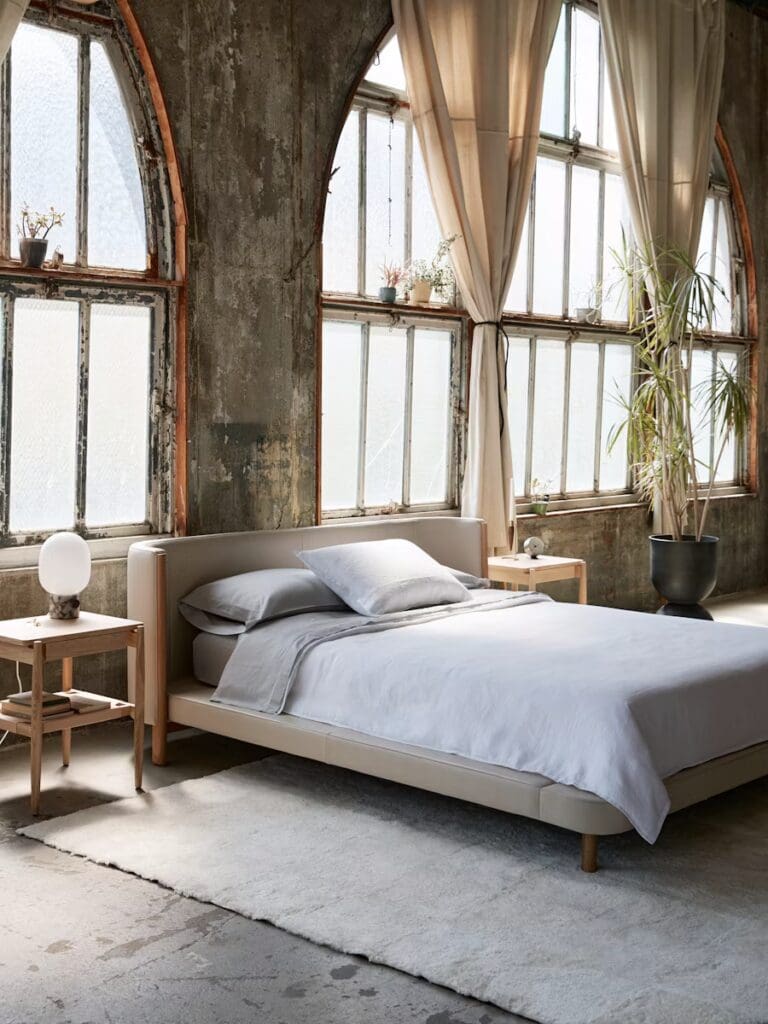 A modern bedroom featuring a light-colored bed with a minimalist design, soft bedding, and wooden side tables. Large arched windows with sheer curtains allow natural light to fill the space, complemented by potted plants.