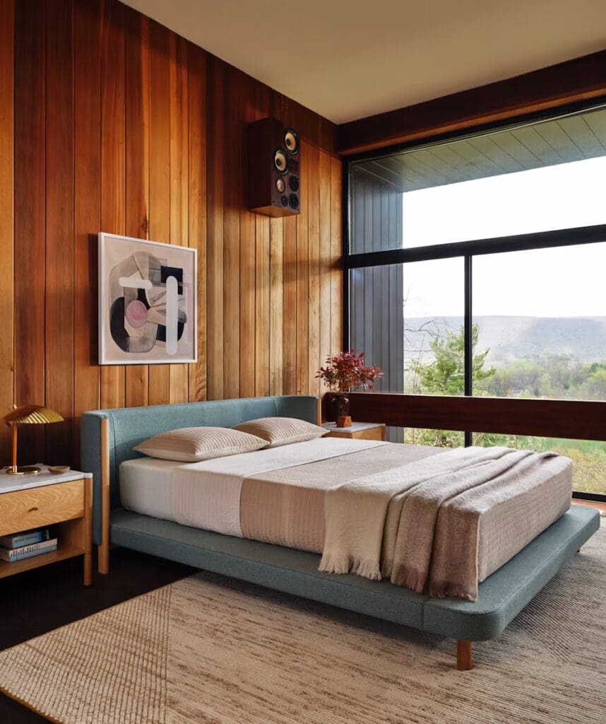A modern bedroom with a wooden accent wall, featuring a light blue bed, soft pillows, a cozy throw blanket, a bedside table with a lamp, and large windows showcasing a scenic view.