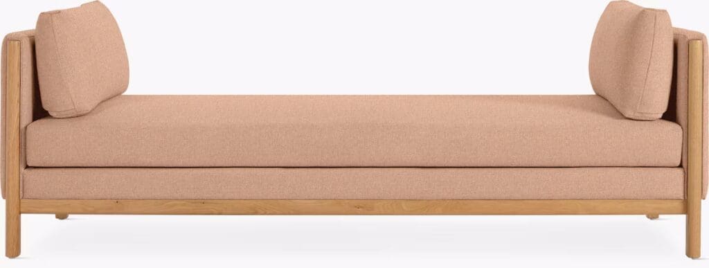 A modern, minimalist daybed with a light pink fabric cushion and wooden frame, featuring two matching back cushions.