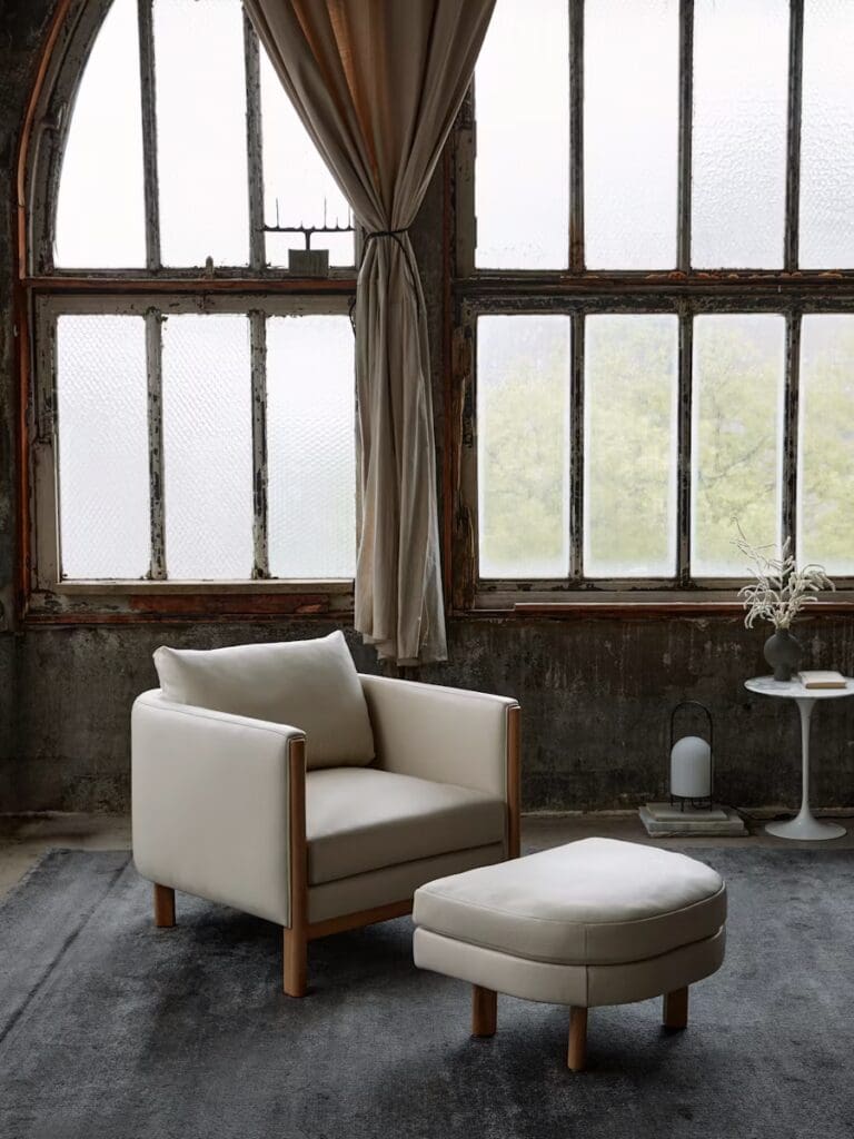 A modern living room featuring a cream-colored armchair with wooden accents and a coordinating ottoman, set against large industrial-style windows with curtains. A side table with a decorative item is visible.