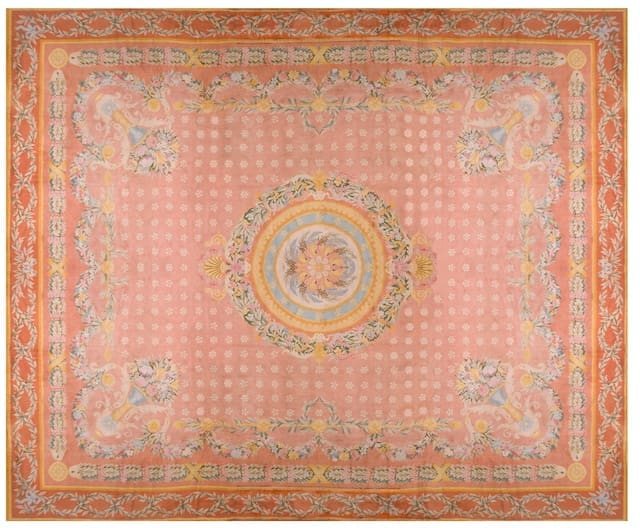 A vintage rug with intricate floral patterns in soft pink, orange, and blue tones, featuring a central medallion and ornate border.