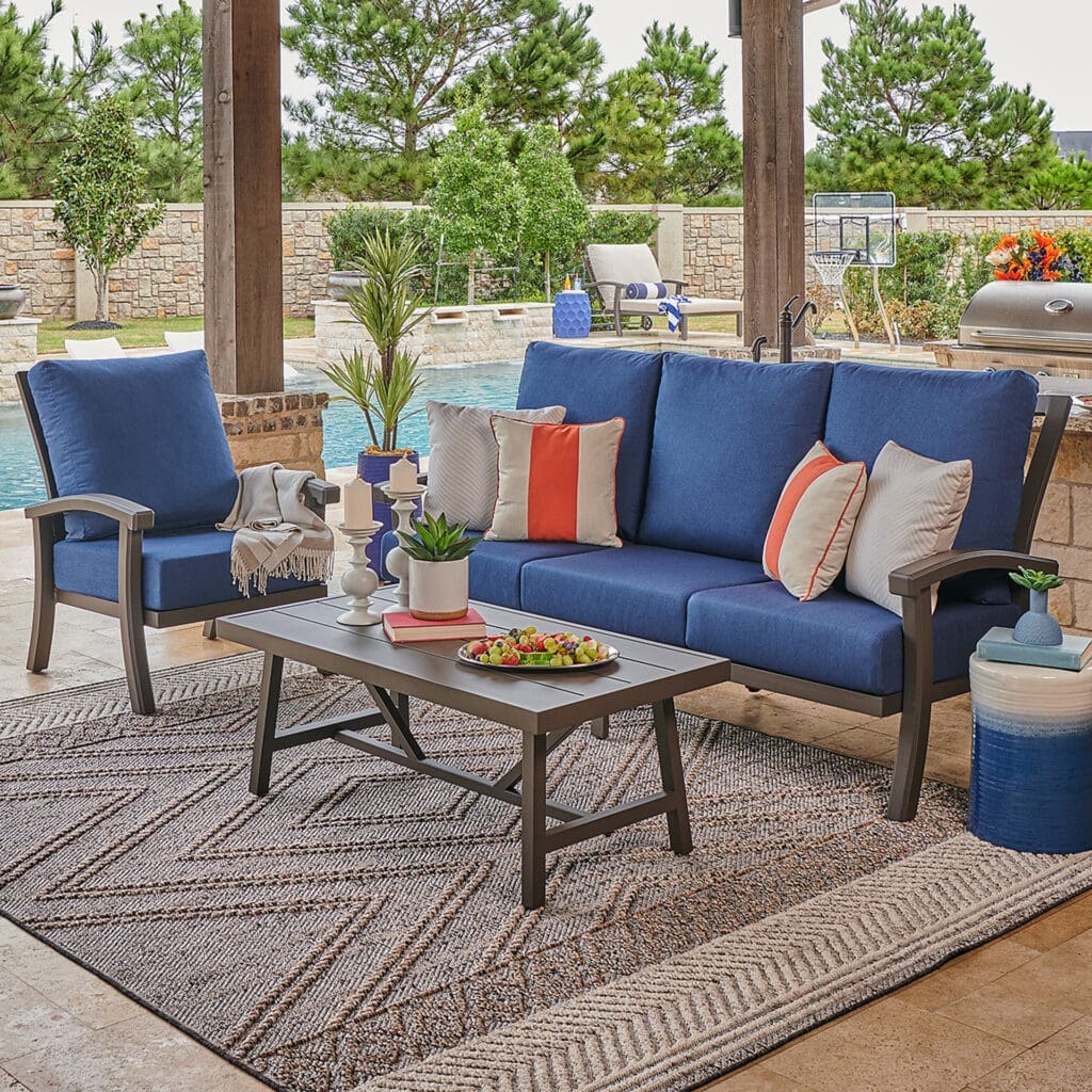 Cozy outdoor living area featuring a blue sofa set with vibrant cushions, a coffee table adorned with snacks, and decorative elements like potted plants, all next to a sparkling pool.