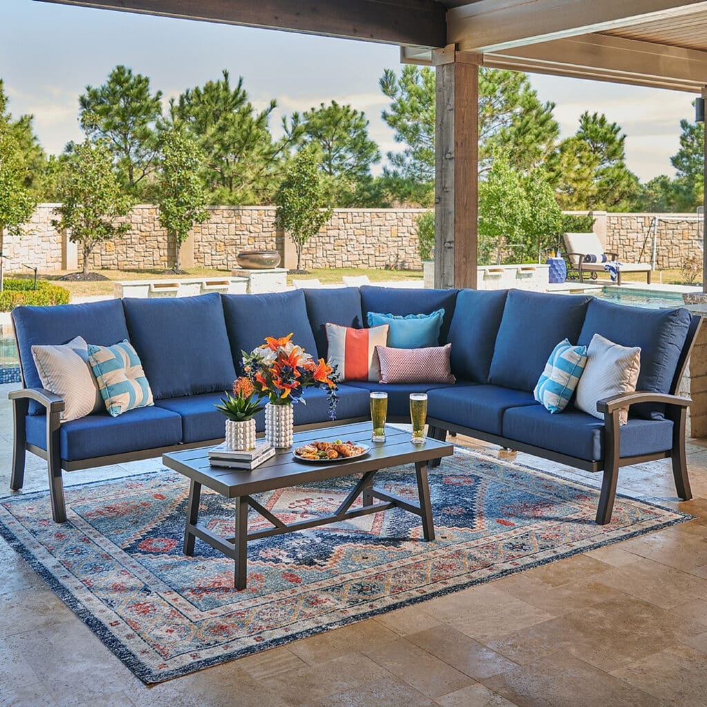 Luxurious outdoor living space with a navy blue sectional sofa, decorated with colorful pillows, and a glass coffee table set with drinks and snacks. The setting features a patterned area rug and lush greenery in the background, suggesting an inviting retreat.