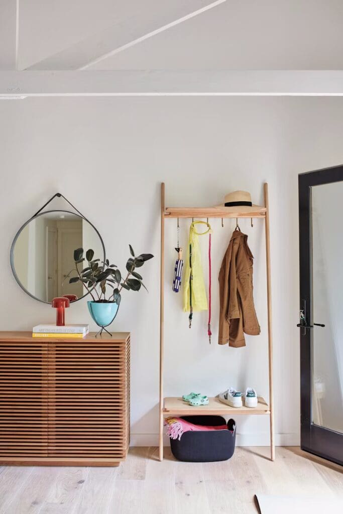 A stylish entryway featuring a wooden coat rack with hanging garments, a round mirror, a wooden console with books, and a potted plant. The floor is light-colored wood and a door is visible in the background.