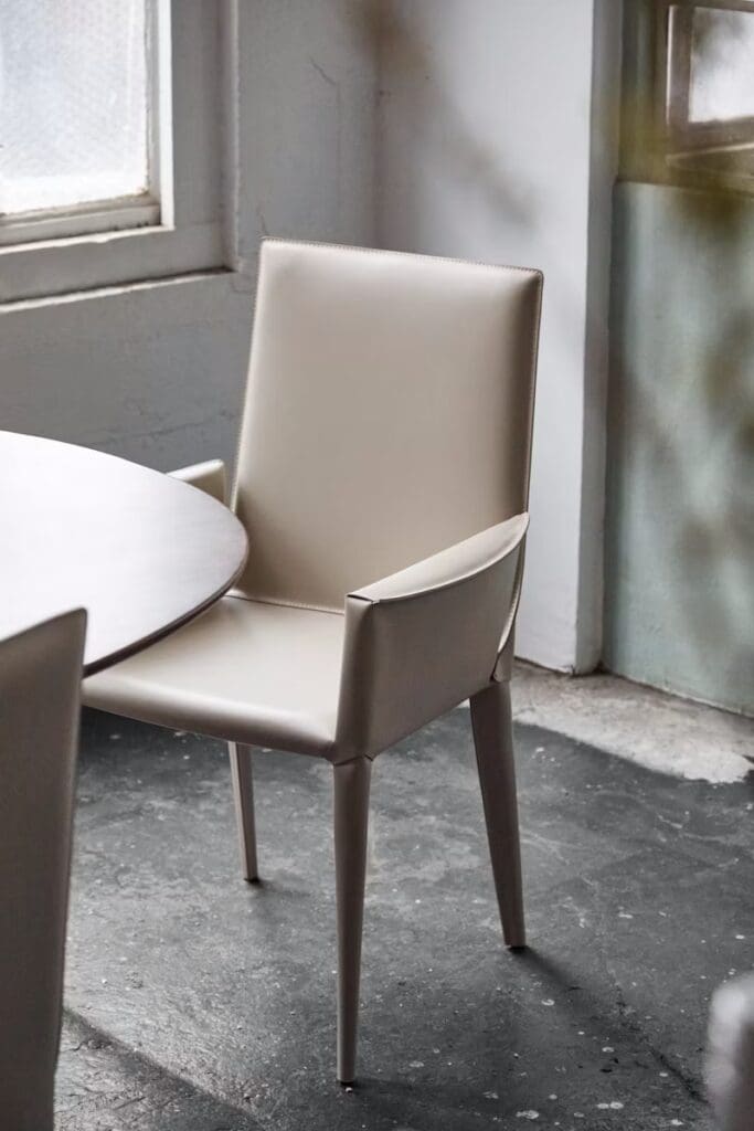 A stylish beige dining chair positioned next to a round table in a modern interior, with a concrete floor and large windows providing natural light.