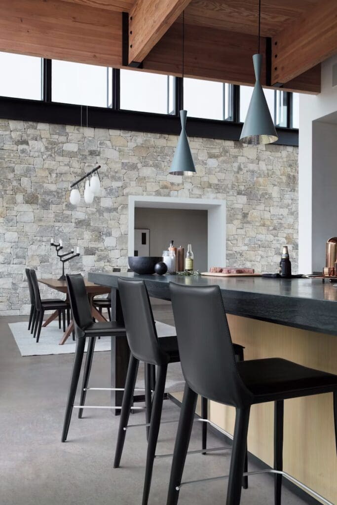 A modern kitchen featuring a stone wall, sleek bar stools, and contemporary pendant lights hanging from a wooden ceiling. The space is open and bright, designed with a minimalist aesthetic.