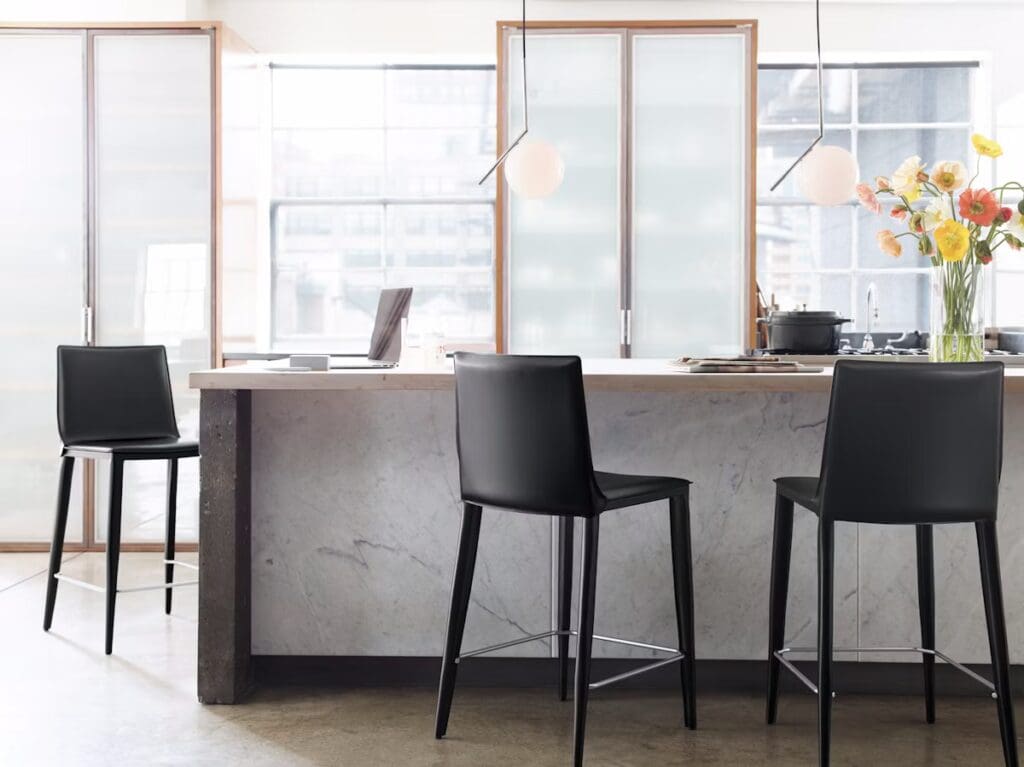 A modern kitchen space featuring a sleek marble-topped island with high black bar stools, large windows allowing natural light, and a vase of vibrant flowers on the counter.