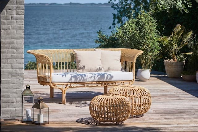 Stylish outdoor seating area featuring a rattan couch with cushions, round wicker tables, and lush greenery, overlooking a serene ocean view.
