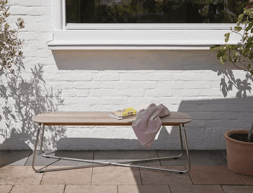 Minimalist outdoor bench with a wooden top and metal legs, set against a white wall. A pink blanket and a book rest on the bench, next to a potted plant.