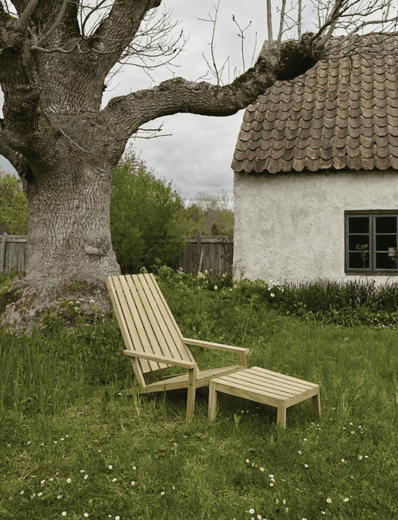 Wooden lounge chair and footrest on grassy area next to a tree and a small white house with a tiled roof under a cloudy sky.