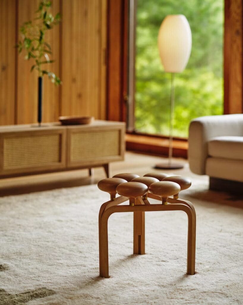 A modern wooden stool with rounded seating elements, placed in a warm living room setting with natural light coming through large windows and a stylish floor lamp in the background.