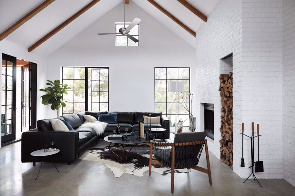 A modern living room with a black sectional sofa, decorative pillows, a wooden coffee table, and a cozy chair. Large windows provide natural light, while a potted plant adds a touch of greenery. A white brick wall with a fireplace and stacked firewood is visible.
