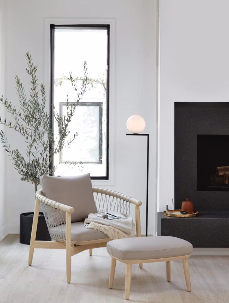 A modern living room featuring a comfortable armchair with an ottoman, a floor lamp, large windows, and a decorative plant.