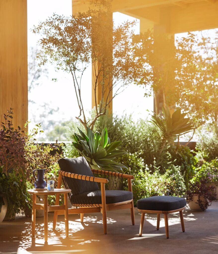 A cozy outdoor seating area featuring a wooden armchair with a dark cushion and a matching footstool, accompanied by a small round wooden table. The space is adorned with lush greenery and potted plants, providing a serene and inviting atmosphere.