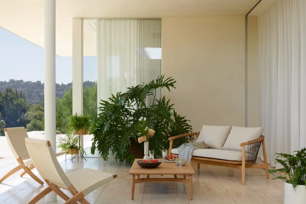 A modern outdoor living area featuring a light-colored sofa with cushions, a wooden coffee table, and two stylish woven chairs. The space is adorned with lush green plants and large glass windows that offer a view of the landscape.