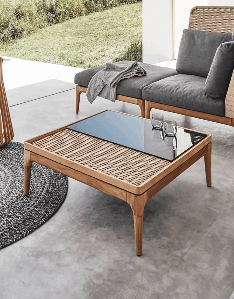 Modern outdoor living area featuring a stylish lounge chair, a woven coffee table, and a round rug on a concrete floor. The backdrop includes a lush green landscape.