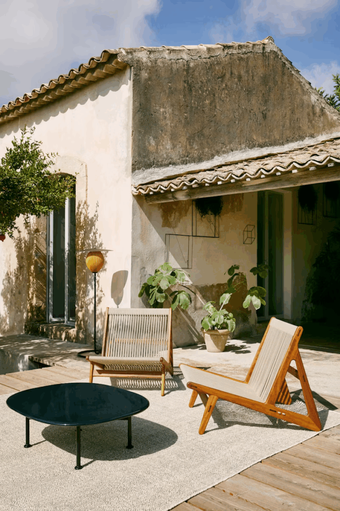 Chic outdoor living area featuring modern wooden furniture, a black coffee table, and potted plants against a backdrop of a rustic house and blue sky.