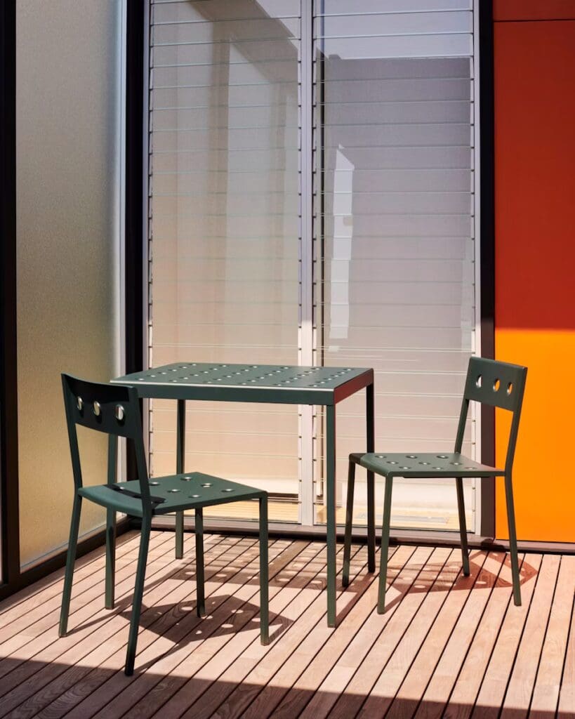 A green outdoor dining set featuring a rectangular table and two matching chairs, placed on a wooden deck with a backdrop of large glass windows.