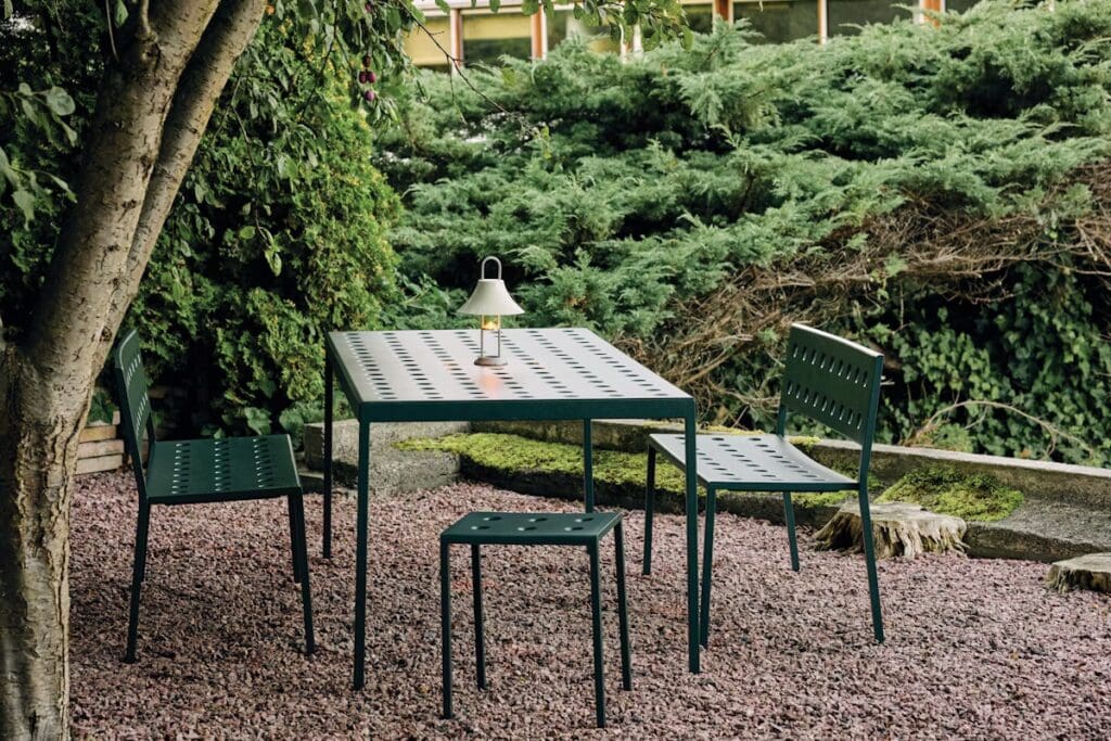 A modern outdoor dining set featuring a green table and matching chairs, surrounded by lush greenery and gravel.