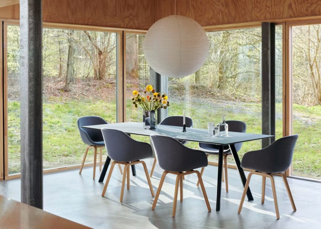 A modern dining room featuring a table with a vase of flowers, surrounded by six stylish chairs, with large windows offering a view of greenery outside.
