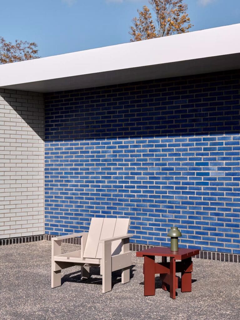 Modern outdoor seating area featuring a light-colored armchair and a red side table with a green lamp, set against a blue brick wall.