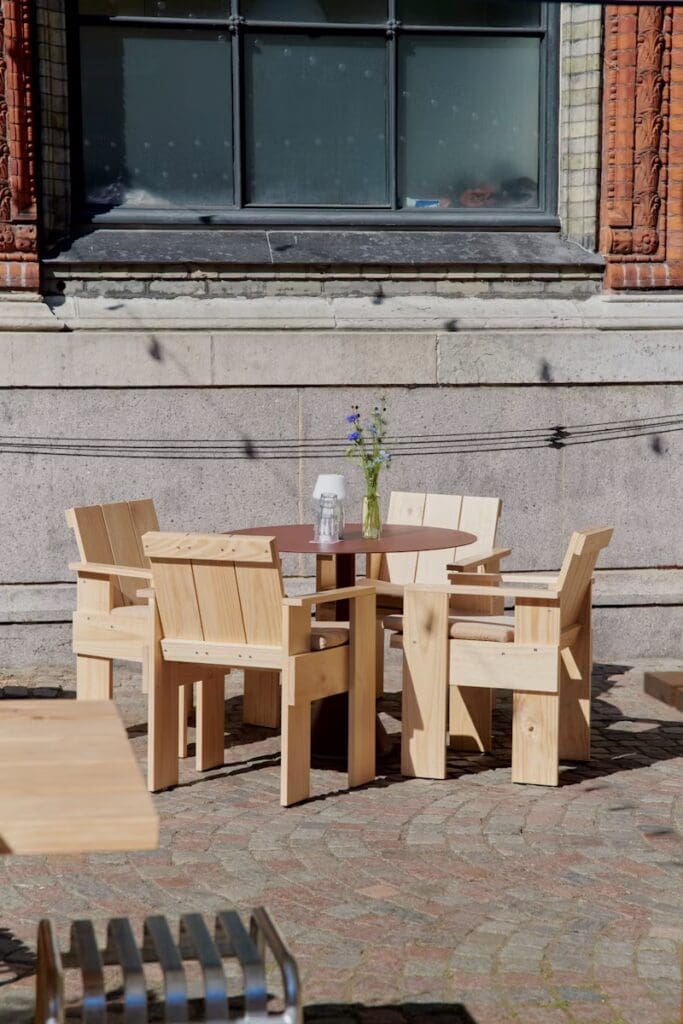 A wooden outdoor dining set featuring a round table and four matching chairs, displayed on a cobblestone patio near a building with decorative brickwork.