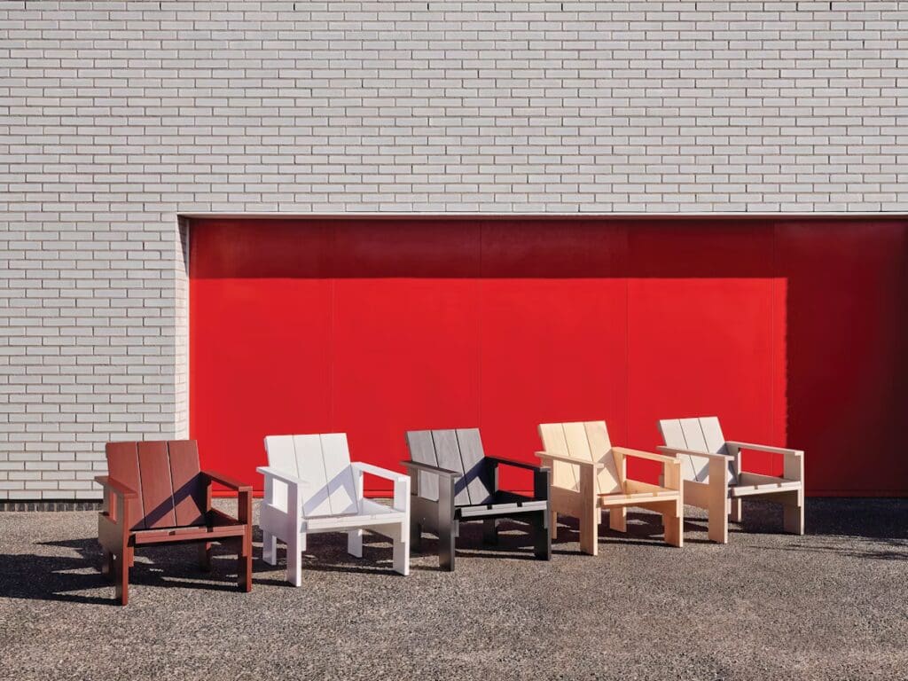 A row of five stylish outdoor chairs in various colors, including brown, white, black, beige, and light gray, positioned in front of a bright red wall.