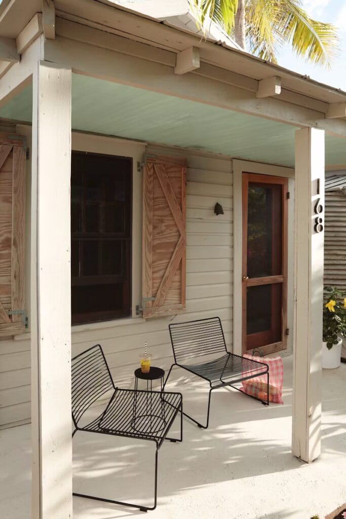 A cozy outdoor seating area featuring two black metal chairs and a small round table with a drink on it, set on a light-colored porch with a wooden door and shuttered windows.