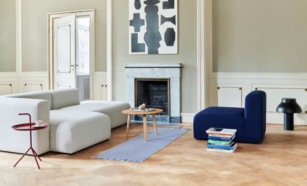 A modern living room featuring a light-colored sectional sofa, a dark blue chair, and a small round table with a cup and books, complemented by a decorative rug and a fireplace.