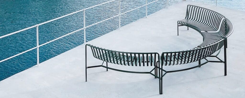 Curved black metal outdoor bench positioned near a body of water, showcasing a modern design with slatted seating.