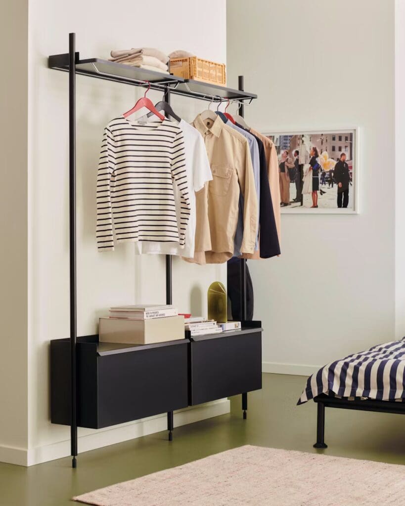 A modern bedroom scene featuring a clothing rack with various shirts, a shelf with a basket, and stylish black storage drawers beneath. In the background, there is a bed with a striped bedspread and a framed photo on the wall.