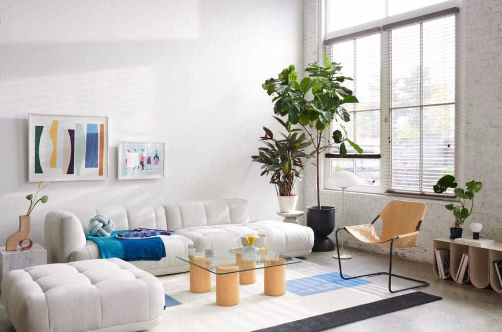 A modern living room featuring a light-colored sectional sofa, decorative pillows, a glass coffee table with wooden legs, and an oversized ottoman. There are plants near the windows and colorful abstract artwork on the walls.
