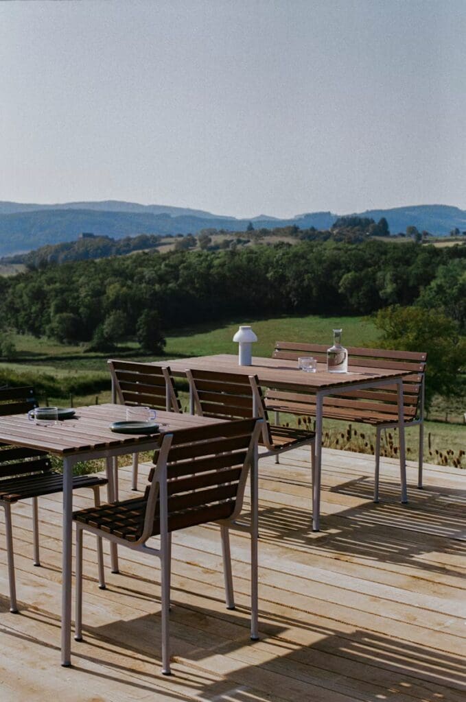 An outdoor terrace with wooden tables and chairs, overlooking a scenic landscape of rolling hills and greenery.