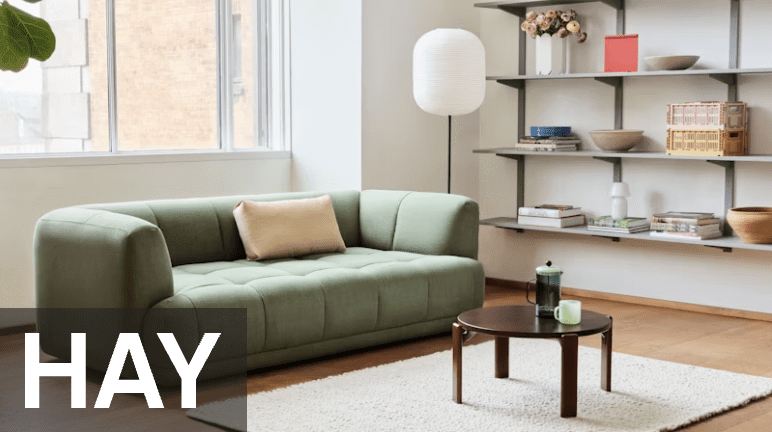 A modern living room with a green sofa, a round coffee table, and a shelf displaying various decor items. Large windows provide natural light.