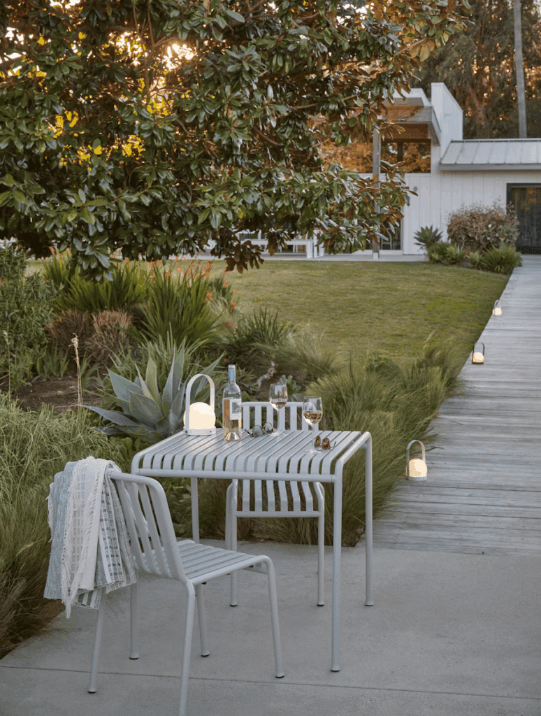 Cozy outdoor dining area featuring a modern table and chairs, with a bottle of wine and glasses placed on the table, surrounded by lush greenery and soft lighting.