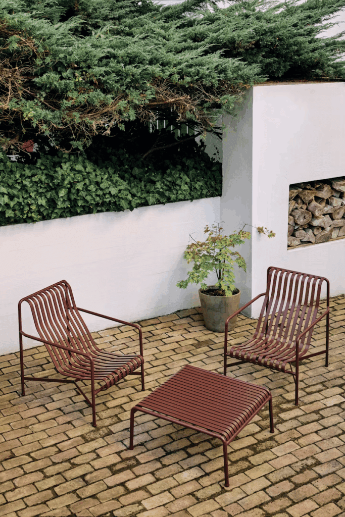 Modern outdoor seating area featuring two red slatted chairs and a slatted coffee table on a brick patio, surrounded by greenery and a white wall.