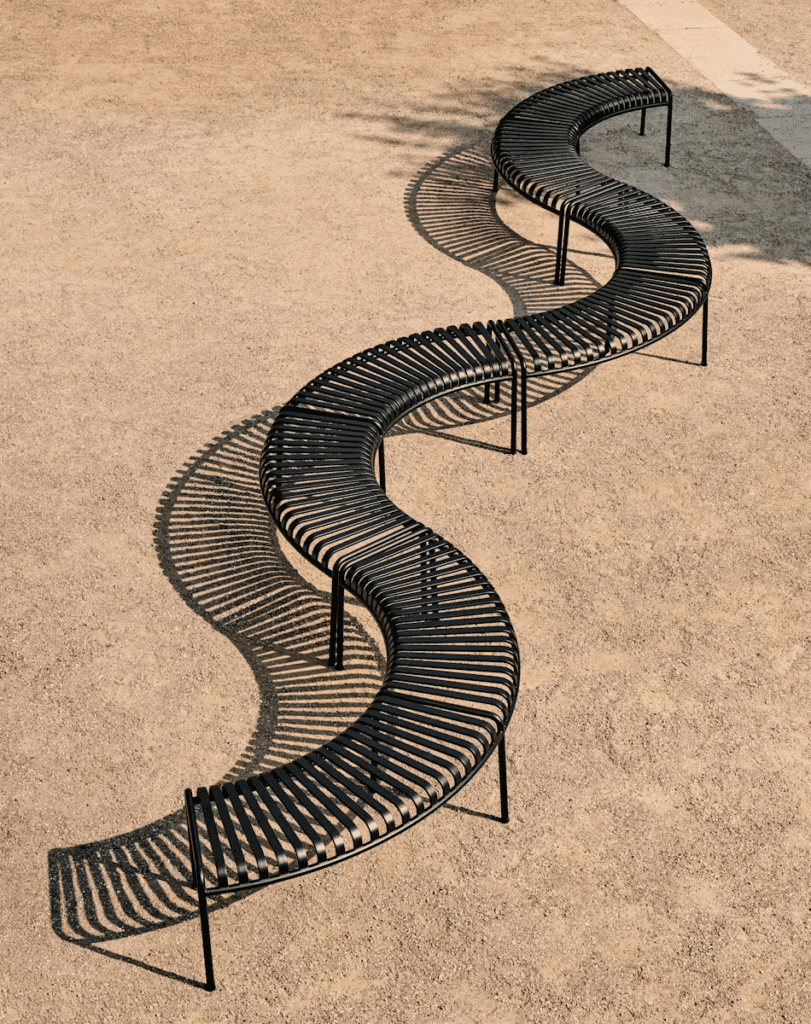 Curved black bench with slotted design placed on a sandy surface, casting intricate shadows.