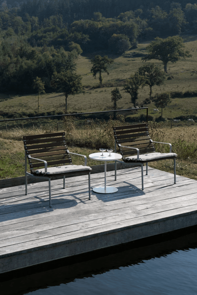 Cozy outdoor seating area with wooden chairs and a small round table, overlooking a serene landscape of trees and fields.