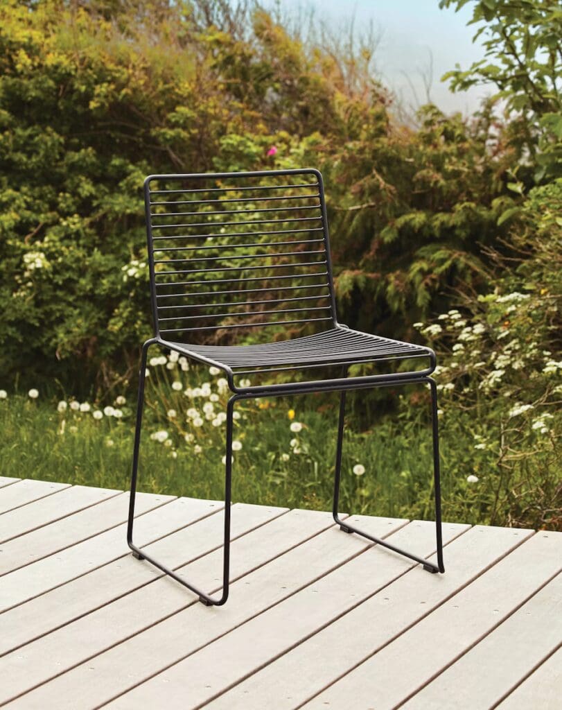 A modern black wire chair placed on a wooden deck, surrounded by greenery and flowering plants.