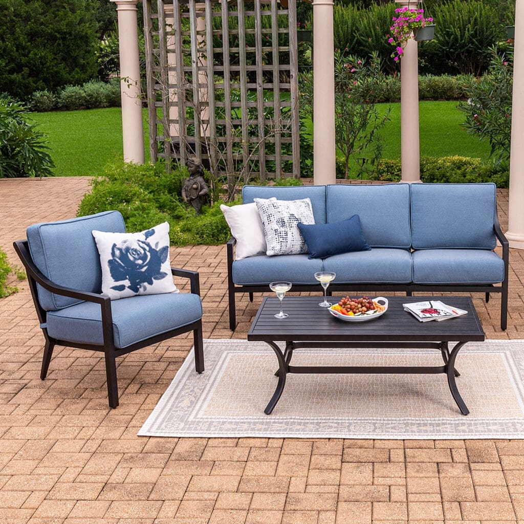 Cozy outdoor seating area featuring a blue sofa and armchair with decorative cushions, a coffee table with snacks and drinks, surrounded by greenery.