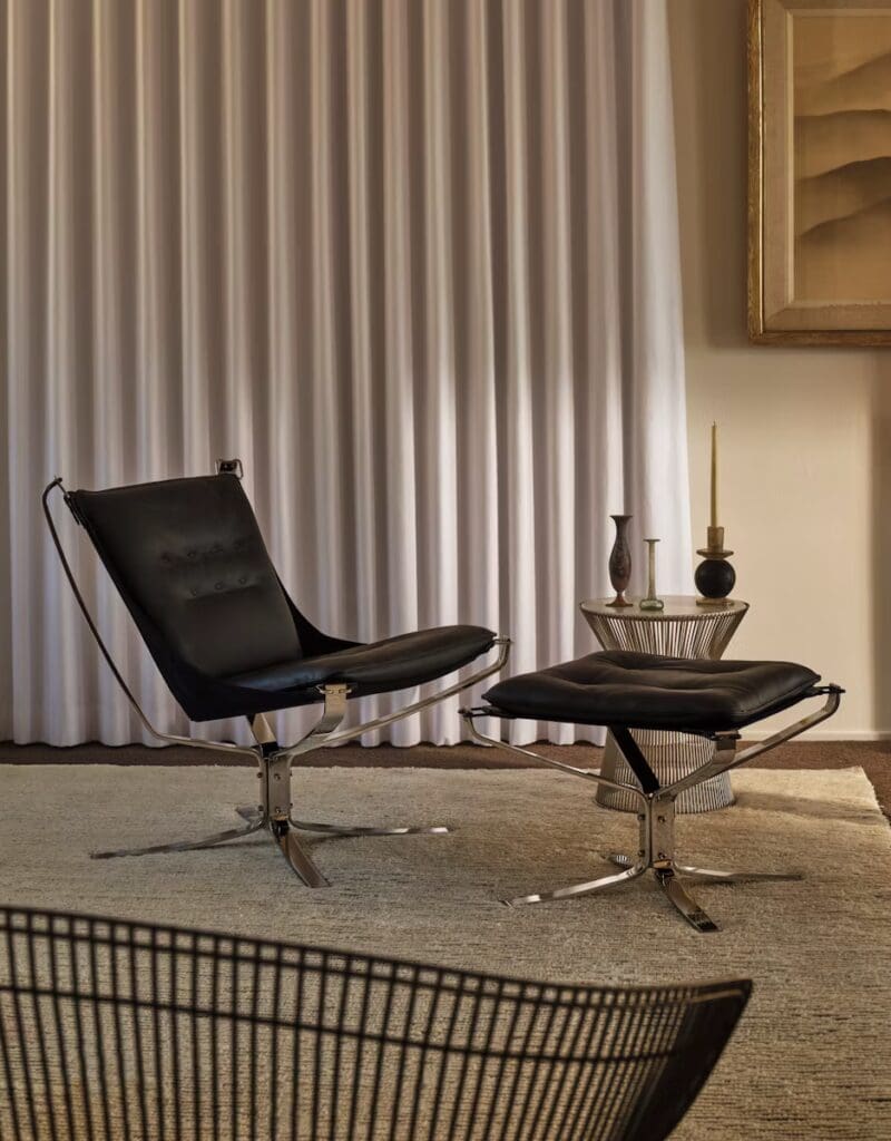 A modern living room with a black lounge chair and ottoman, alongside a decorative side table, set against a backdrop of white curtains and an abstract painting.