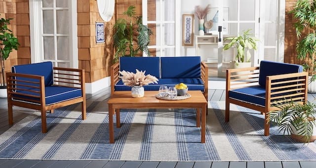 Cozy outdoor living space featuring navy blue furniture and wooden accents, with a coffee table and decorative accessories, set against a light-colored backdrop and greenery.