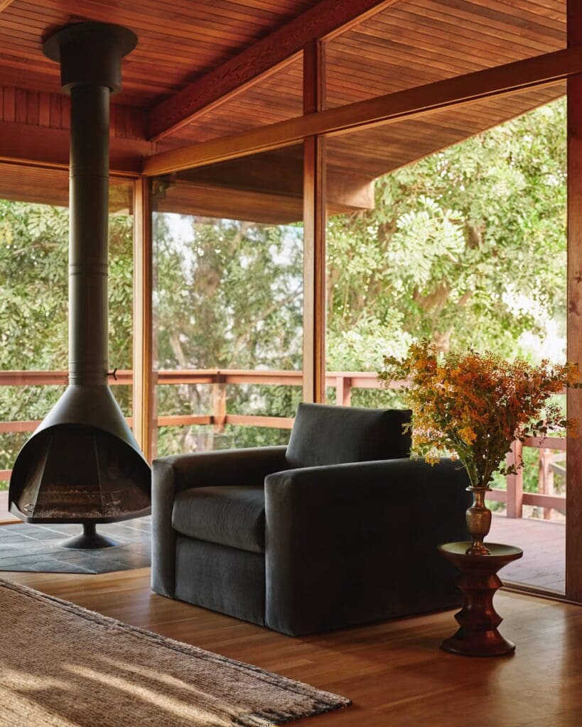 A modern living room featuring a dark armchair, a unique fireplace, and large windows overlooking greenery. A wooden side table holds a vase with flowers, complementing the warm wooden floors and accents.