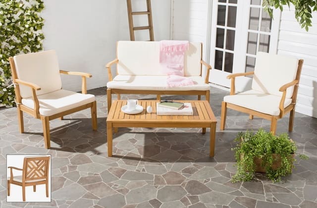 Modern outdoor seating set with cream cushions, featuring two armchairs, a loveseat, and a coffee table, set on a stone patio with greenery in the background.