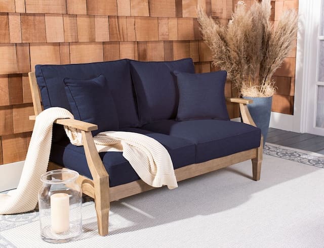 Cozy outdoor sofa with navy cushions and wooden armrests, complemented by a beige throw blanket and decorative plants in the background.