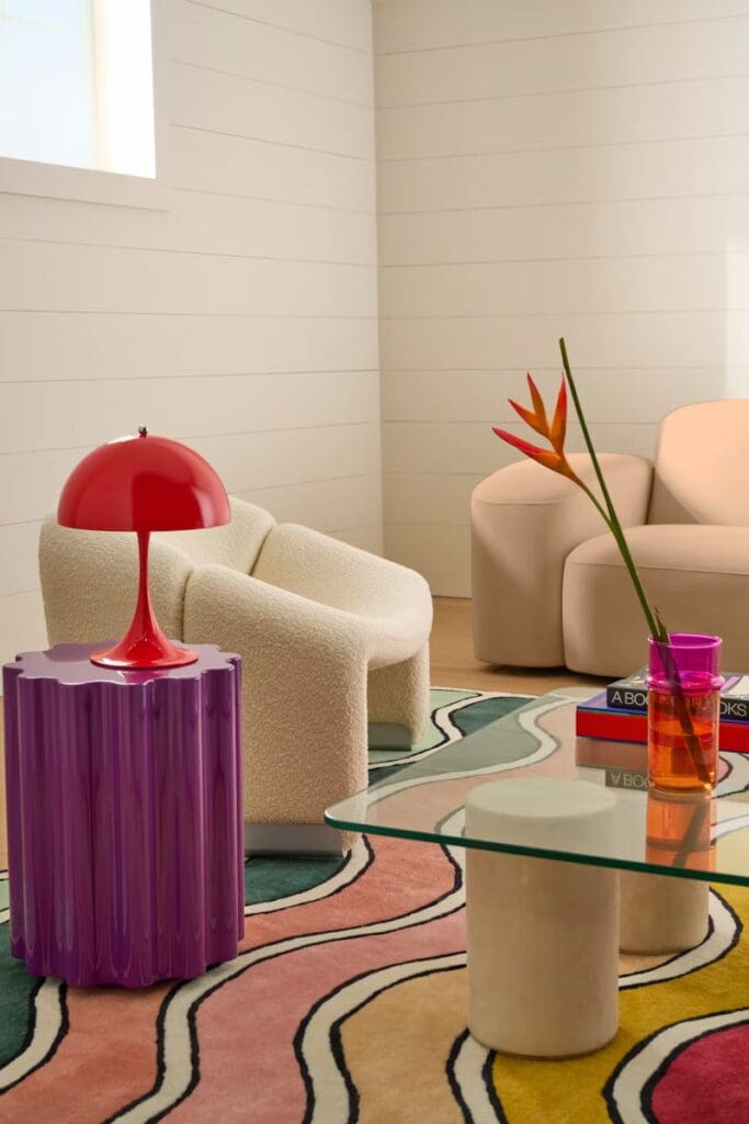 A vibrant modern living room featuring a large colorful rug, a glass coffee table, a purple side table with a red lamp, and a plush armchair with a textured fabric.
