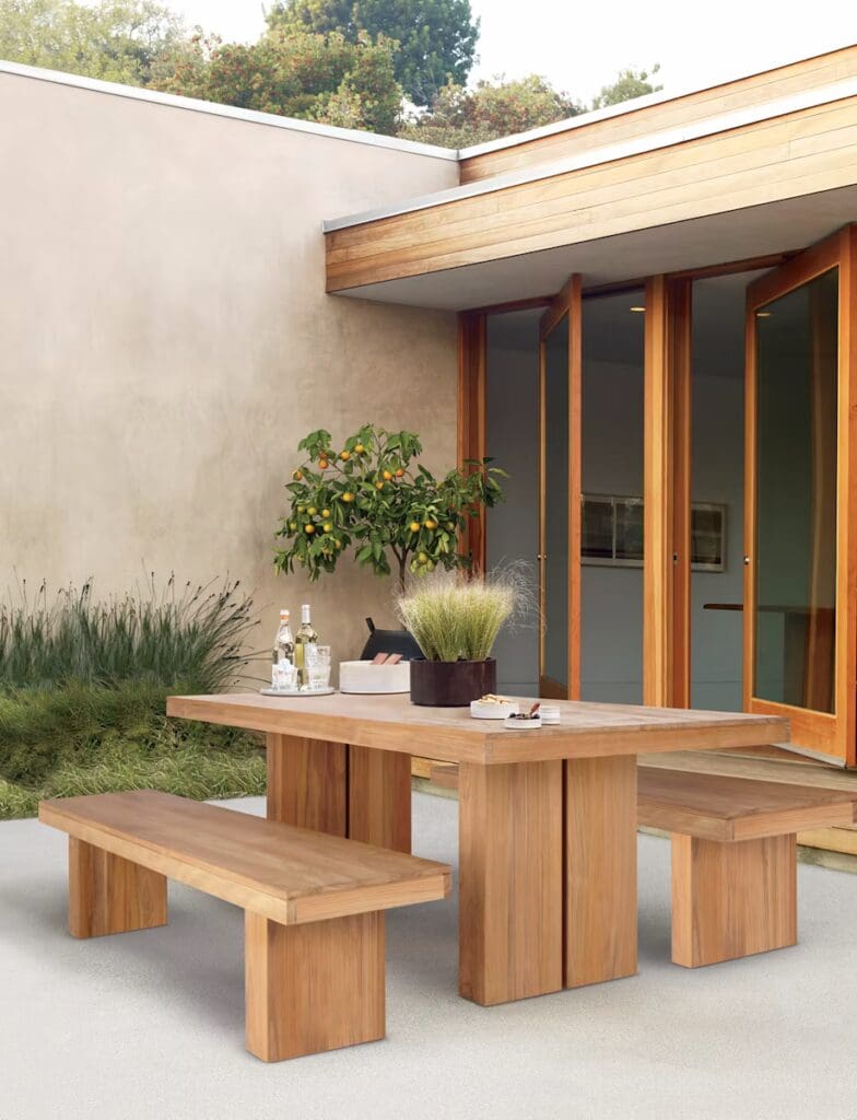 An outdoor dining area featuring a wooden table with matching benches, a small plant centerpiece, bottles of wine, and a modern architectural background.