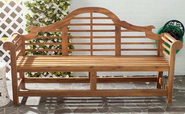 Wooden garden bench with a decorative backrest and slatted seat, positioned against a white wall and surrounded by greenery.